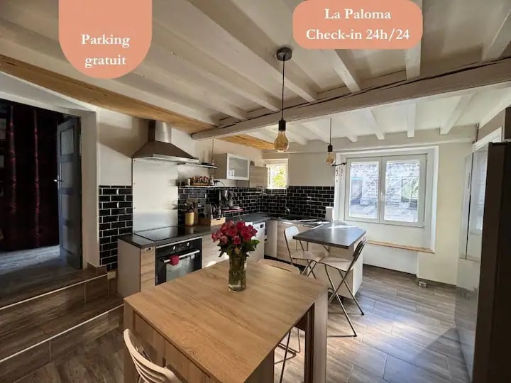 A modern kitchen is presented with sleek appliances and a central wooden dining table. Bright natural light fills the space, highlighting the black tiled backsplash and hanging light fixtures. Fresh flowers are placed on the table, adding a touch of color to the functional layout.