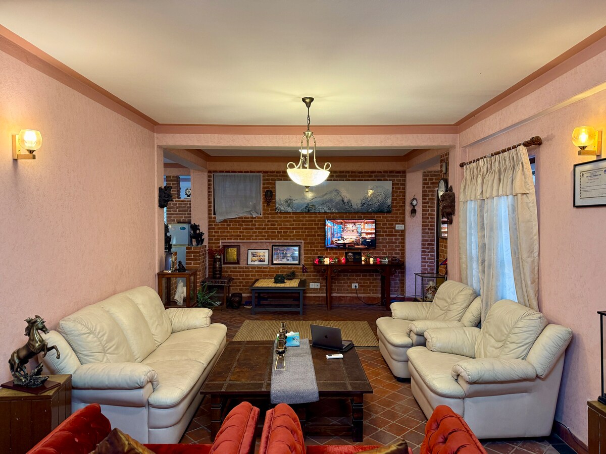 A spacious living area features soft leather sofas arranged around a large wooden coffee table. The room is accented by warm lighting and a classic chandelier. A television is mounted on the wall, with decorative items displayed on shelves beneath it. Large curtains frame the window, providing natural light.