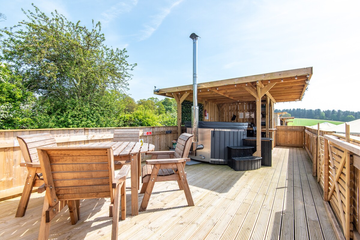 An elevated decking area is featured, surrounded by greenery. A wooden dining table with chairs is positioned in the foreground. A private hot tub is located next to a wooden shelter, providing options for relaxation while enjoying the outdoors.