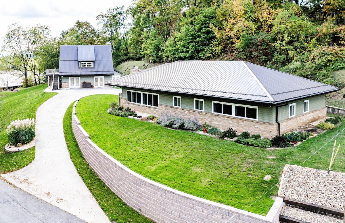 The exterior view of a modern ranch-style home is captured, featuring a well-maintained lawn with landscaped gardens. A paved drive leads to the home, which displays large windows on its facade. Surrounding greenery enhances the serene and inviting atmosphere of the property.