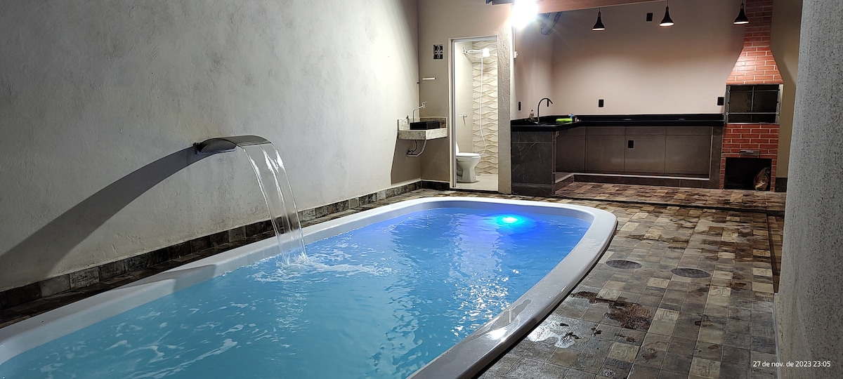 An indoor swimming pool is featured, with a gentle waterfall flowing into it. Soft lighting casts a serene glow over the water. A kitchenette area and a barbecue grill are visible in the background, with modern finishes and tiled flooring enhancing the space.