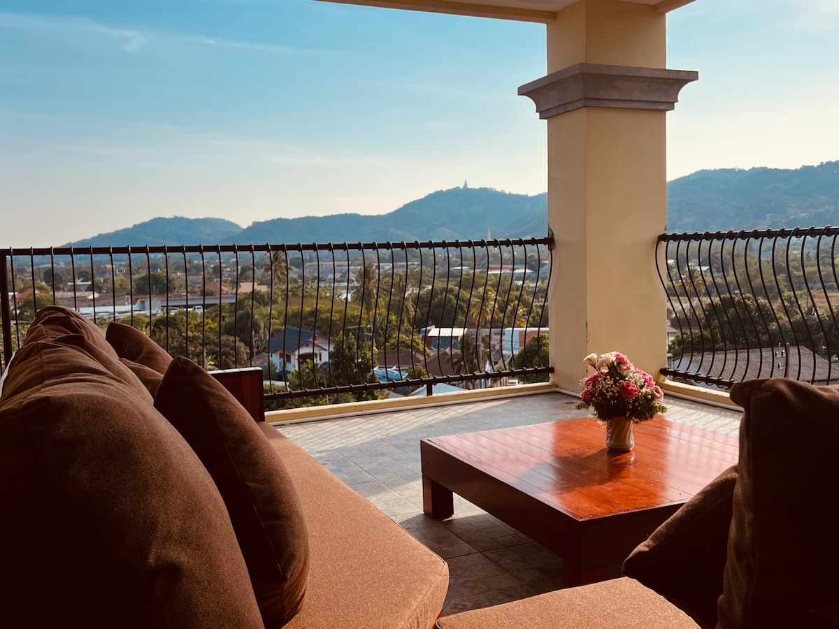 A generous balcony is furnished with large, plush seating and a wooden coffee table, offering a scenic view of rolling hills and distant landscapes. A floral arrangement adds a touch of charm, while the expansive space invites relaxation under a clear sky.