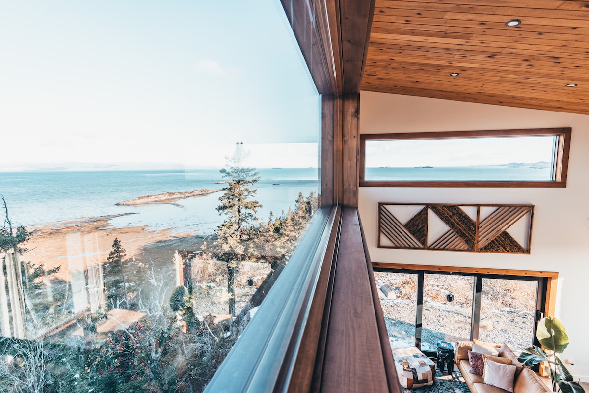 A panoramic view of the river is captured through large windows, highlighting the serene water against a backdrop of trees and distant islands. Natural light fills the space, while contemporary wooden accents create a warm setting within the living area.
