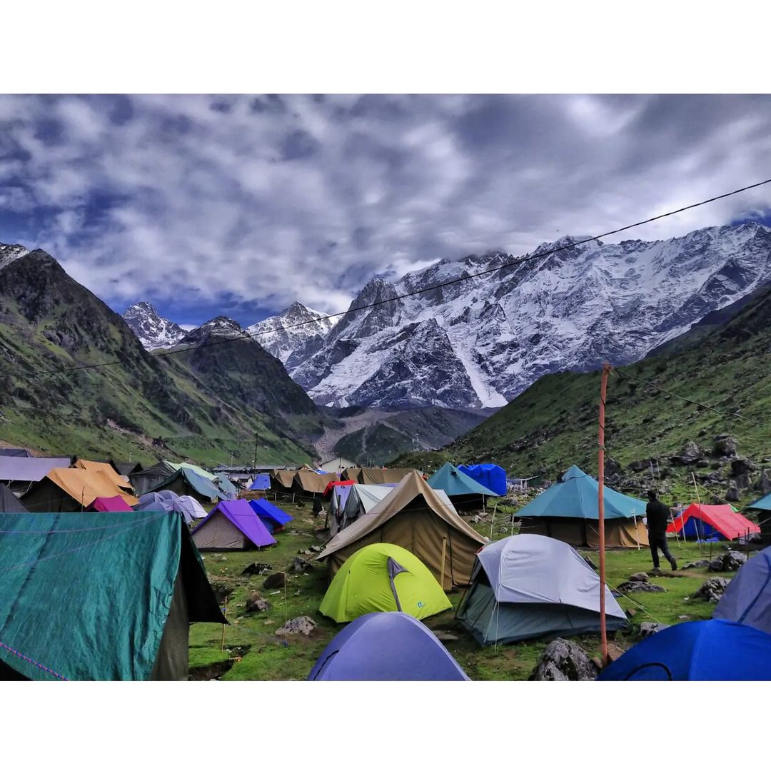A scenic view of multiple colorful tents arranged in a mountainous area, surrounded by lush green grass and rocky terrain. Majestic snow-capped peaks rise in the background under a mostly cloudy sky, creating a peaceful camping atmosphere.