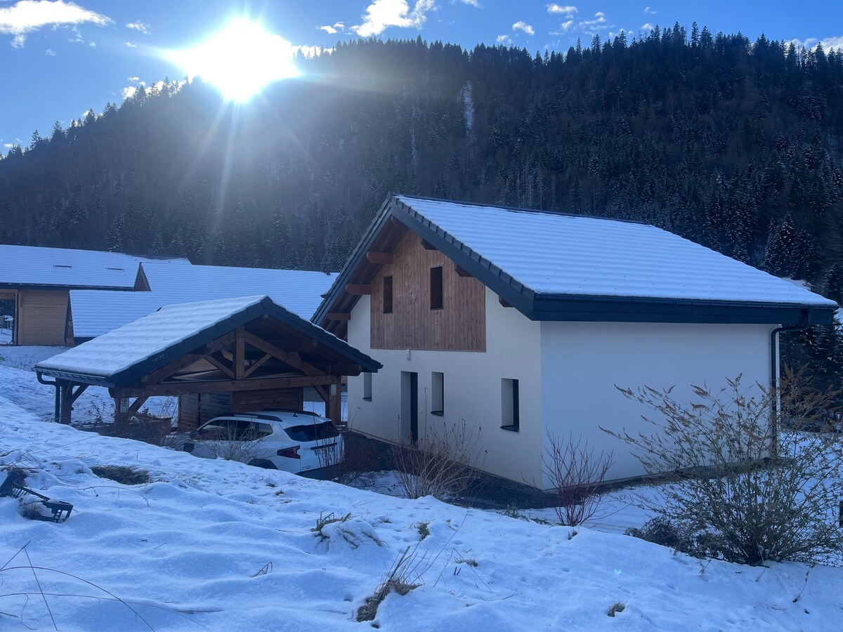 A newly constructed chalet is nestled amidst snow-covered landscapes, featuring a sloped roof and wooden accents. A spacious carport is visible alongside the building, with the sun shining brightly in the background, casting warm light over the serene winter scene.
