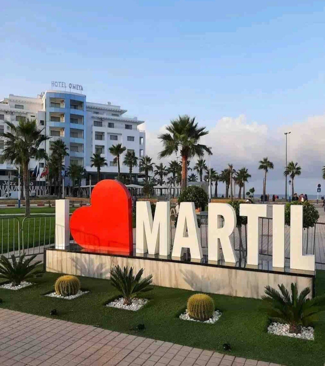 The image captures a large, vibrant 'I ❤️ MARTIL' sign set against a backdrop of a modern hotel. Surrounding the sign, well-maintained greenery includes palm trees and decorative cacti, while a paved walkway provides access to the scenic area nearby.