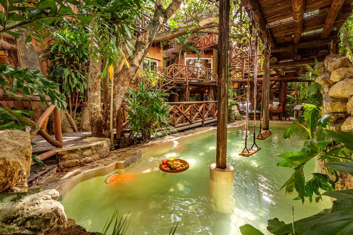 A serene swimming area, inspired by a natural cenote, is surrounded by lush greenery and wooden structures. Hammocks hang invitingly above the calm water, which reflects the vibrant foliage. The tranquil atmosphere is complemented by natural rock formations, offering a peaceful retreat amidst nature.