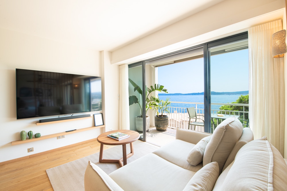 A spacious living area features a large QLED TV mounted on the wall. A comfortable sofa is positioned adjacent to floor-to-ceiling glass doors that open to a balcony. Indoor plants add a refreshing touch, while the stunning view of the sea is prominently visible.