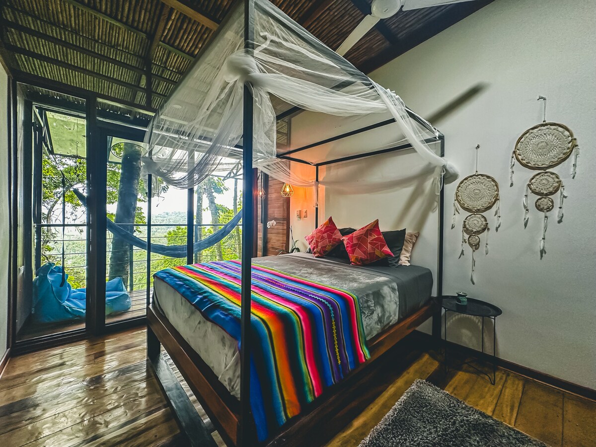 A spacious bedroom features a four-poster bed with a colorful striped blanket. Large windows offer natural light and views of the surrounding jungle. Dreamcatchers adorn the wall, and a cozy hammock can be seen outside, inviting relaxation amid nature.