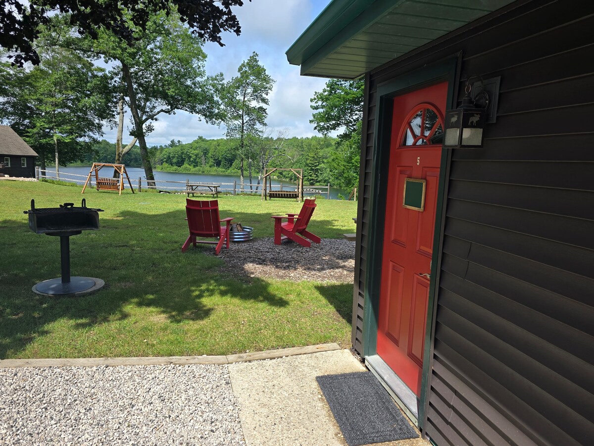 A vibrant red door provides entry to the cabin, framed by a gravel path. Just outside, colorful chairs are arranged around a fire pit. Lush green grass leads to the lake, with trees and a swing set visible in the background.