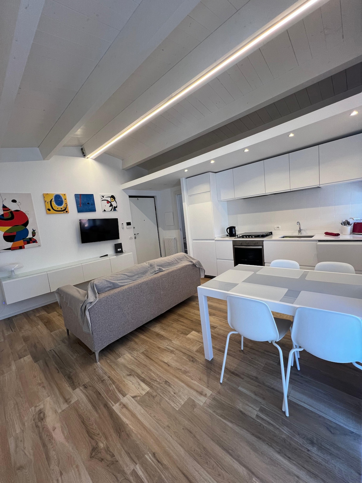 A spacious living area featuring a beige sofa positioned by a wall-mounted television. A dining table with white chairs is located near the kitchen, which includes modern appliances and ample cabinet space. Bright, neutral tones complement the light wood flooring.