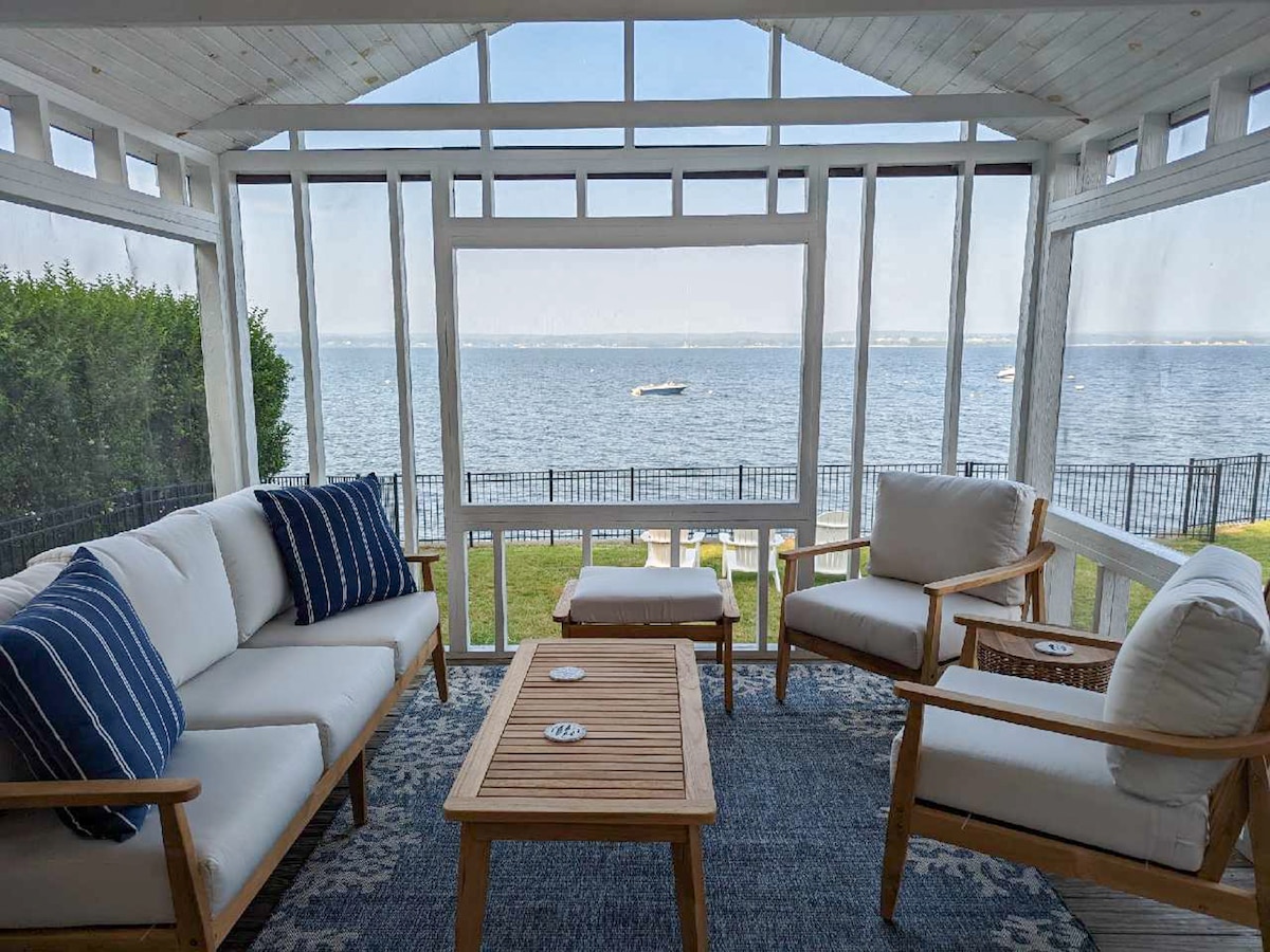 A screened-in porch provides unobstructed views of the water, featuring comfortable seating arranged around a wooden coffee table. Natural light floods the space, highlighting the tranquil scenery outside.