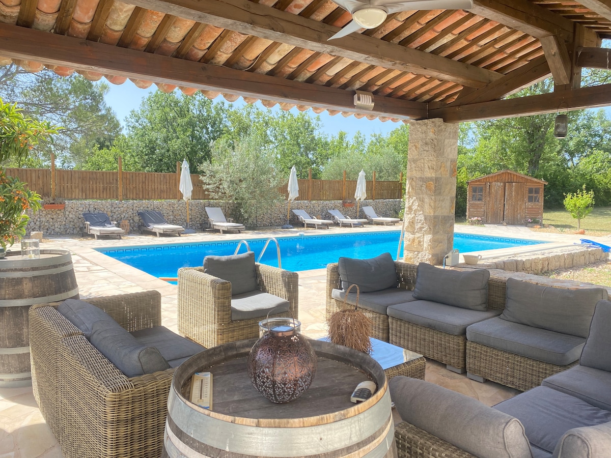 A shaded outdoor seating area features comfortable grey sofas arranged around a wooden coffee table. A stone-walled pool is visible in the background, accompanied by sun loungers and umbrellas. Lush greenery and a wooden shed are also present, contributing to a serene atmosphere.