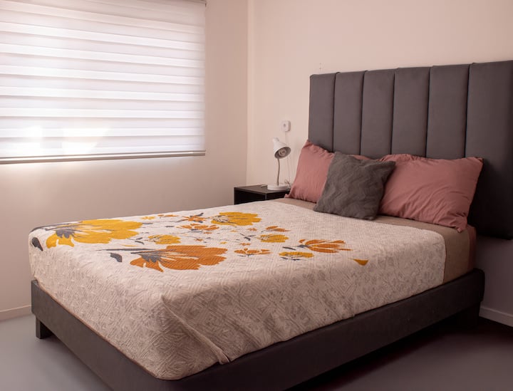Quarto confortável e luminoso com uma cama grande, cabeceira acolchoada e mesa lateral com lâmpada para um descanso relaxante.