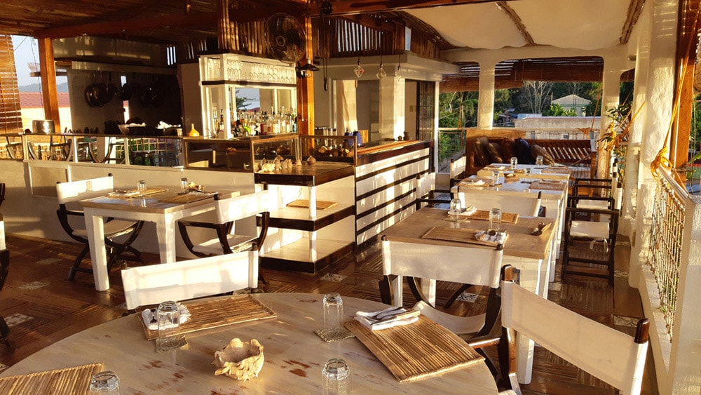 An Italian restaurant features an open-air design with wooden accents and a spacious floor plan. Tables are arranged with place settings, and a bar is visible in the background. Natural light filters through, illuminating the inviting dining area and surrounding greenery.