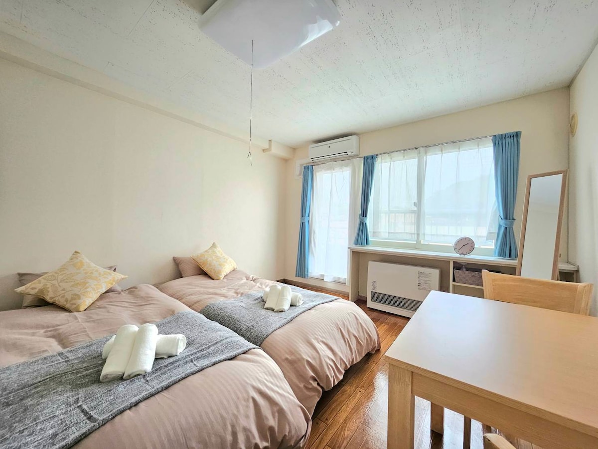 A well-lit bedroom features two spacious beds adorned with soft blankets and decorative pillows. A wooden table is positioned nearby, complemented by a mirror. Large windows allow natural light to fill the room, with soft blue curtains adding a touch of color.