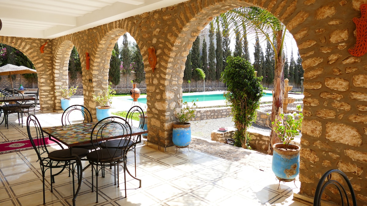 An outdoor terrace is shaded by arches made of natural stone, featuring metal dining tables and chairs. Potted plants add greenery, while a swimming pool is visible in the background, surrounded by trees and a well-maintained garden.