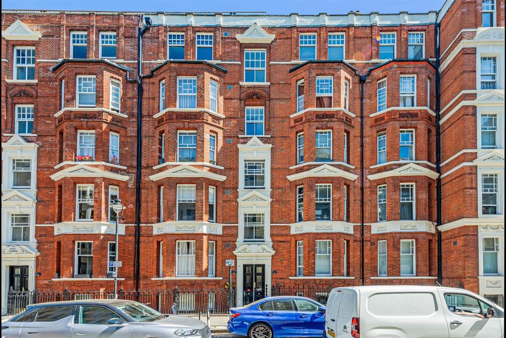 A red brick apartment building with intricate architectural details is featured. The facade showcases multiple windows and balconies, providing a sense of character. Cars are parked along the street in front of the building, adding to the urban environment.