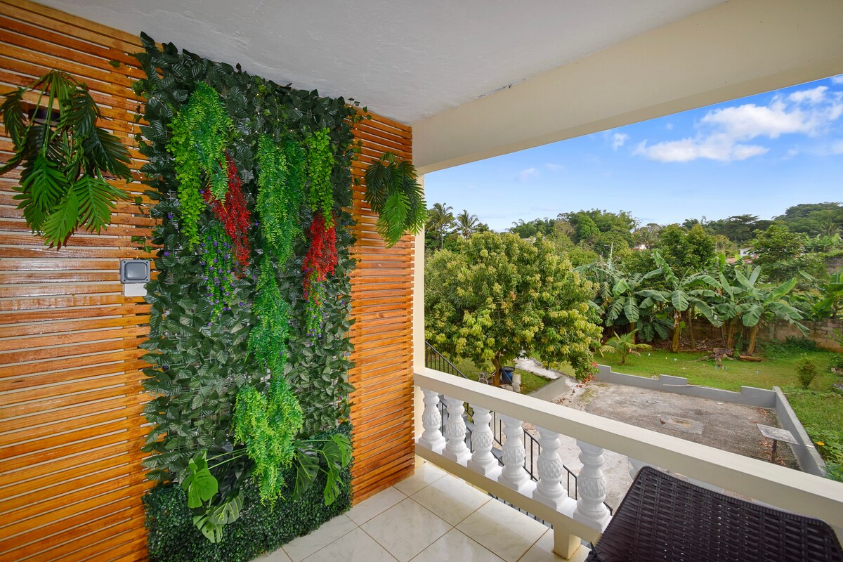 A private balcony is adorned with a vertical garden of vibrant greenery, featuring lush foliage and splashes of red. The view reveals a serene garden area with trees and plants, providing a tranquil backdrop for relaxation among nature.