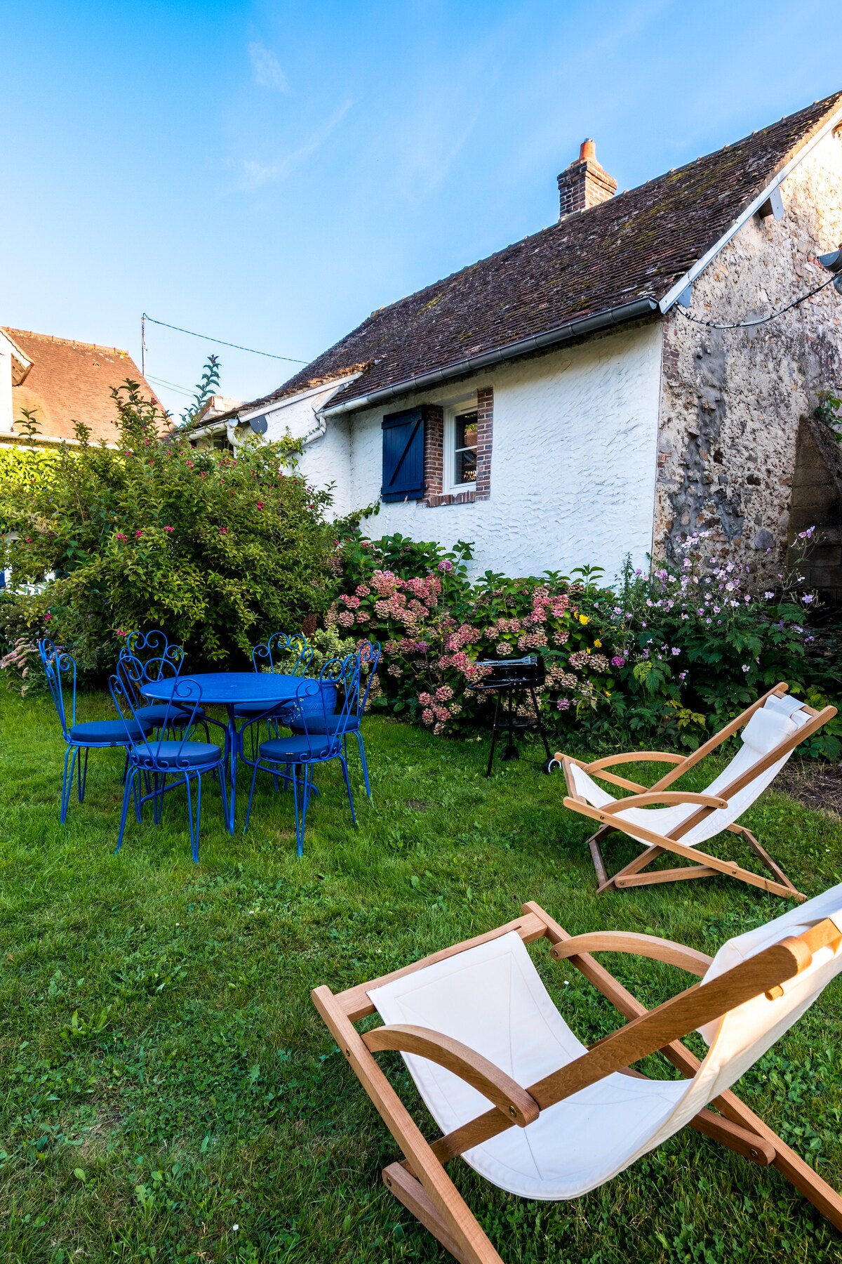 A charming outdoor space is featured, showcasing a blue table surrounded by matching chairs. Two lounge chairs are positioned nearby on the green grass. Lush greenery and flowering shrubs add a natural backdrop to the garden area, accompanied by a barbecue grill for outdoor cooking.