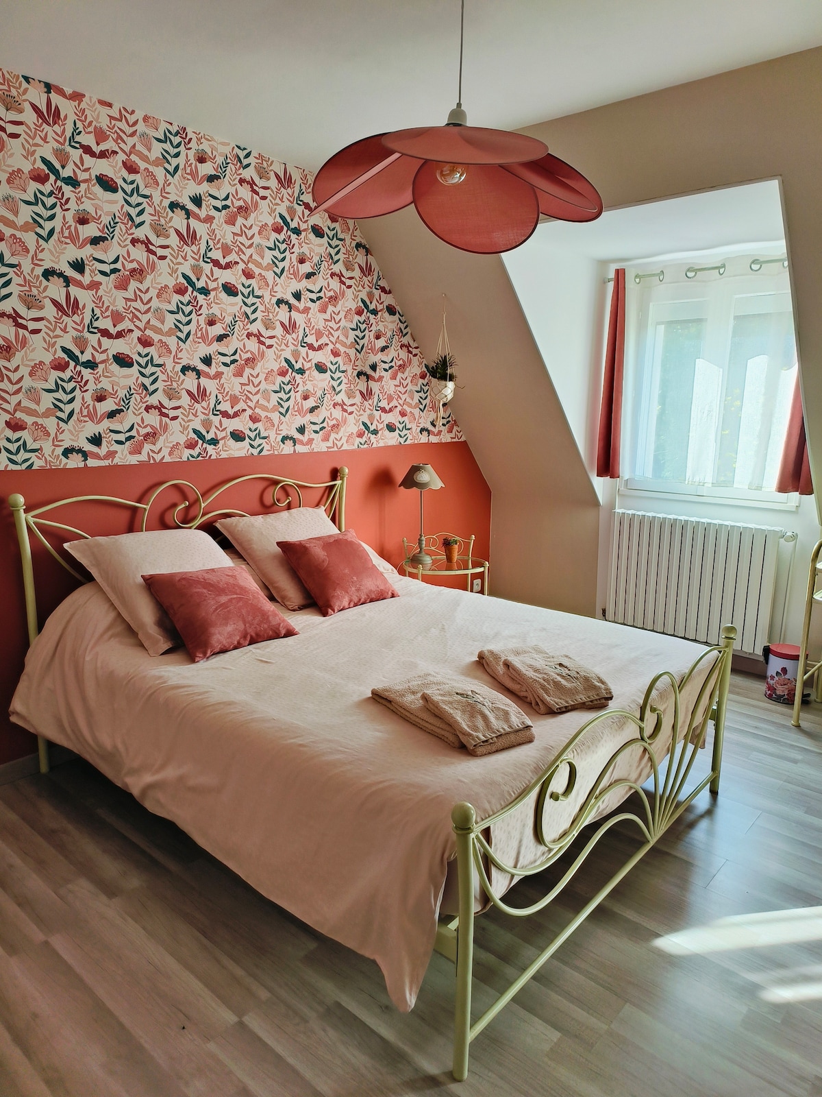 A charming bedroom is featured with a large double bed adorned with soft linens and two decorative pillows. Colorful floral wallpaper adds character to the wall, while natural light streams in through a window covered with light curtains. A unique flower-shaped light fixture enhances the room's ambiance.