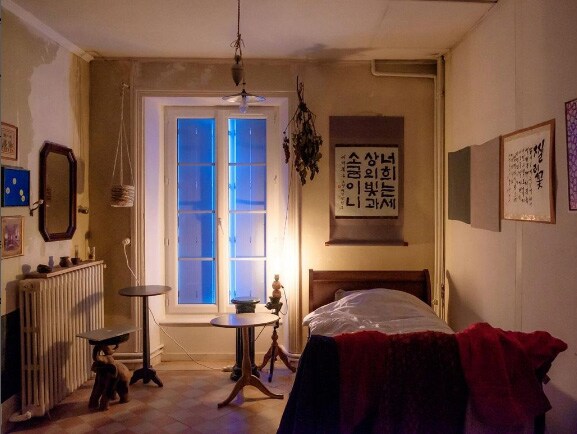 A room with a spacious bed draped in a red blanket is complemented by wooden side tables. A window with a soft blue glow illuminates the space, while framed artwork and a mirror adorn the walls. A radiator and decorative items add character.