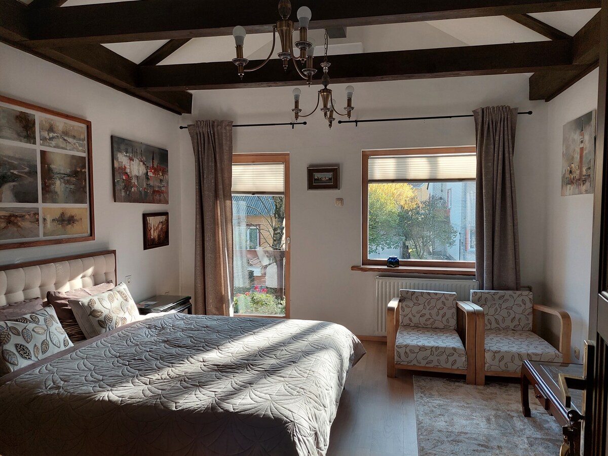 A spacious bedroom features a large bed with a plush headboard, adorned with decorative pillows. Natural light filters through two large windows, illuminating the room. Wooden beams accentuate the ceiling, while a pair of armchairs and a small table provide additional seating.