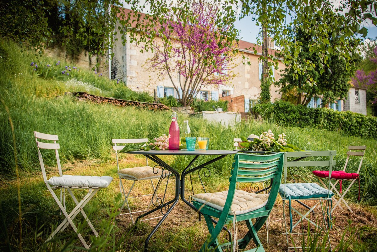 An inviting outdoor seating area is surrounded by lush greenery and blooming flowers. A round table is set with a pink bottle and colorful cups. Four chairs, featuring soft cushions in pastel colors, are positioned around the table, creating a relaxed atmosphere.