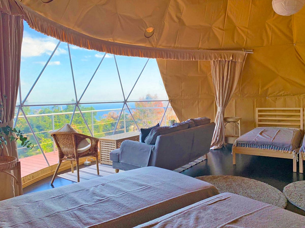 A spacious glamping tent features large windows offering views of the surrounding landscape and ocean. Two single beds are positioned alongside a cozy seating area with a sofa. Natural light fills the room, highlighted by the warm tones of the tent's interior.