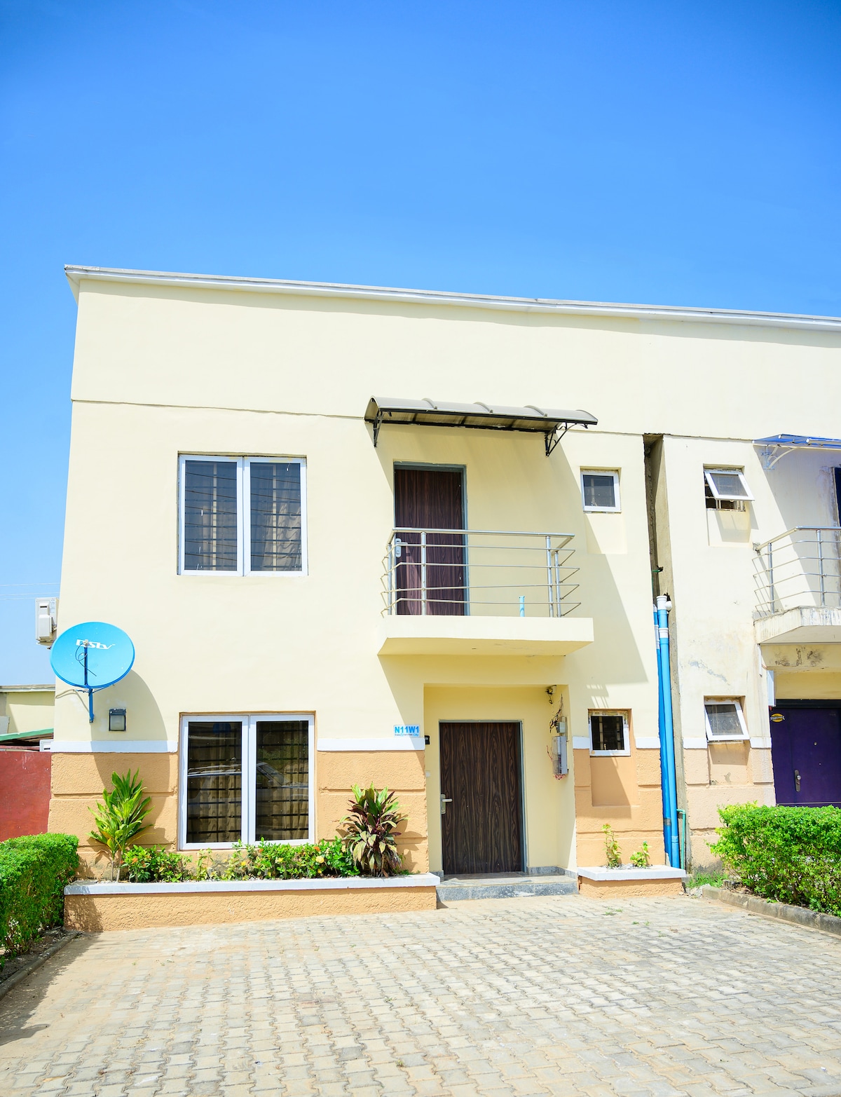 The exterior of the duplex features smooth, cream-colored walls and a front door framed by two large windows. A small balcony extends above, offering a view of the surroundings. A satellite dish is mounted on the wall, and a paved pathway leads to the entrance.