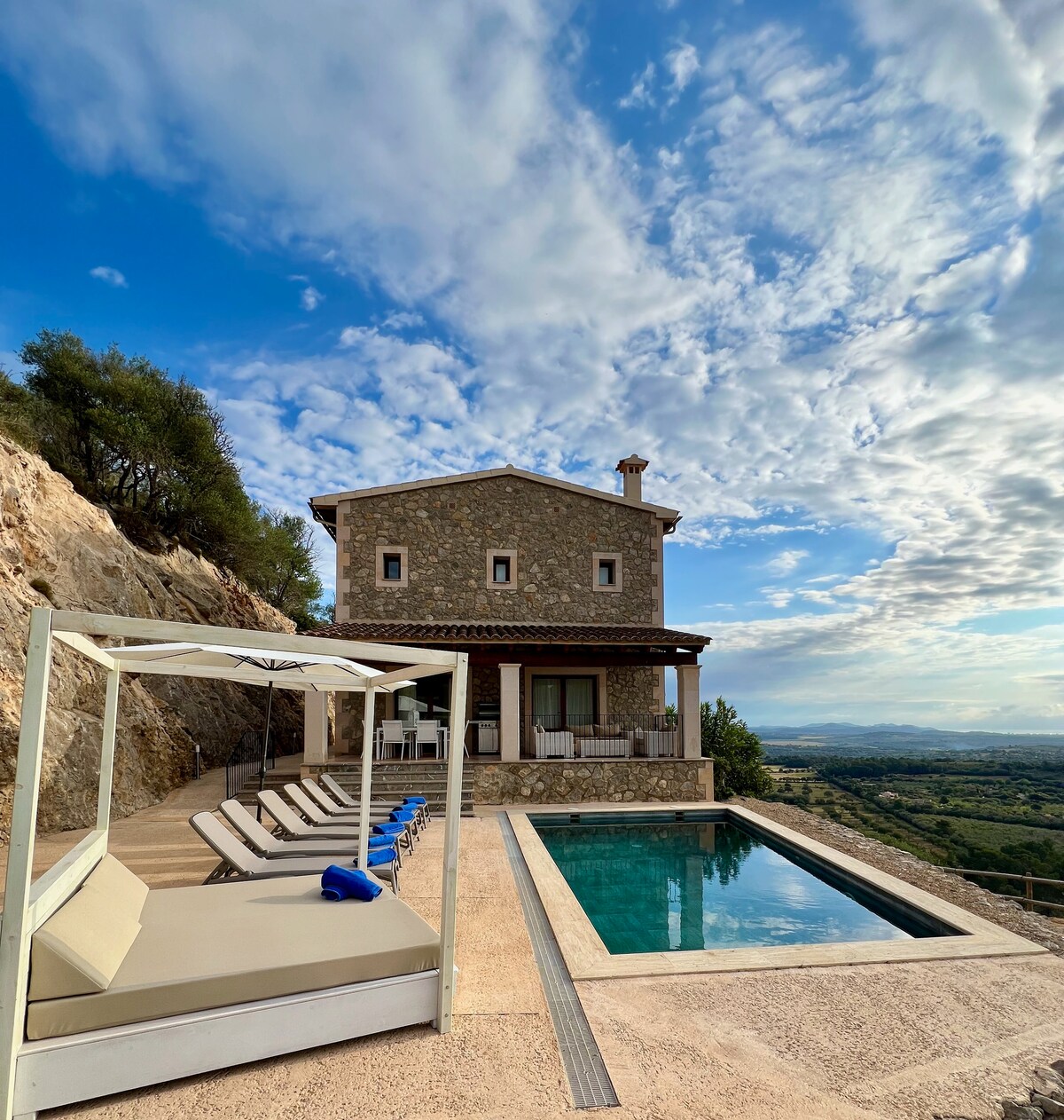 A stone villa is set against a backdrop of rolling hills and a blue sky with scattered clouds. Nearby, a sparkling pool reflects the surroundings, and several loungers are arranged around the pool area, inviting relaxation under the sun.