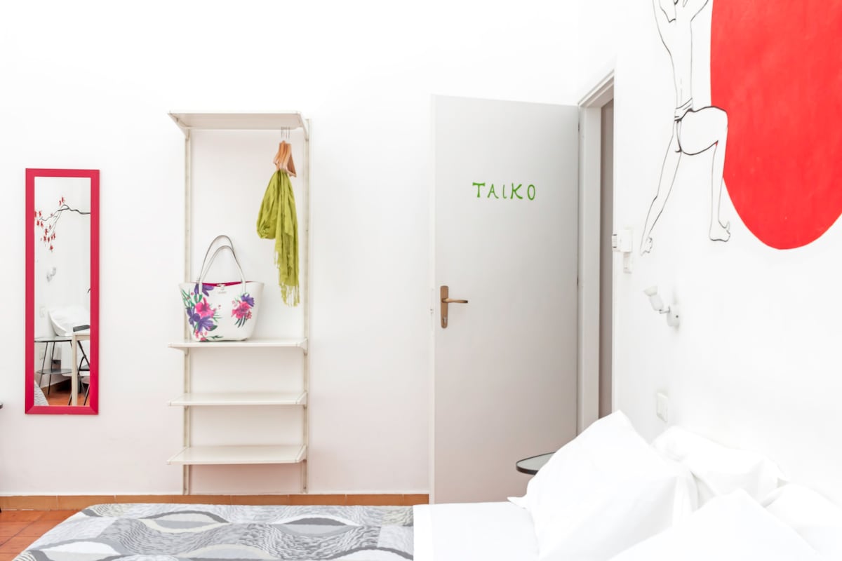 A bright and spacious bedroom is showcased, featuring a large bed with white linens. A minimalistic white shelf holds a handbag and a piece of clothing. A vibrant red accent wall complements a mirror with a decorative frame, while a door labeled 'TALKO' offers a glimpse into the room's layout.