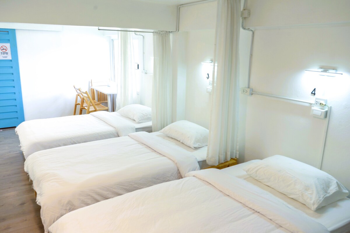 A bright room features three spacious beds dressed in white linens. Soft drapes divide the sleeping area from a small workspace with a table and chairs. Wall-mounted lights provide illumination, while a window allows natural light to fill the space.