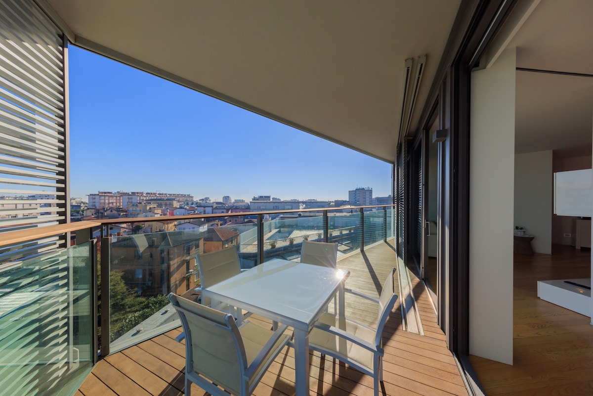An outdoor terrace features a dining table surrounded by several chairs, offering a panoramic view of the surrounding landscape. Glass railings provide an unobstructed sight, while large sliding doors lead into the interior space, enhancing the connection between indoor and outdoor living.