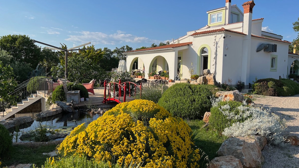 The charming exterior of the property features lush greenery and vibrant yellow flowers. A decorative pond is surrounded by various plants, while a red arched bridge leads to a patio area, providing a welcoming entry to the home.
