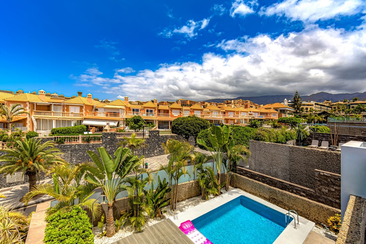 A view of the villa's private heated swimming pool is shown, surrounded by tropical plants and trees. The terrace offers space for relaxation. In the background, other villa structures are visible under a bright blue sky with scattered clouds.