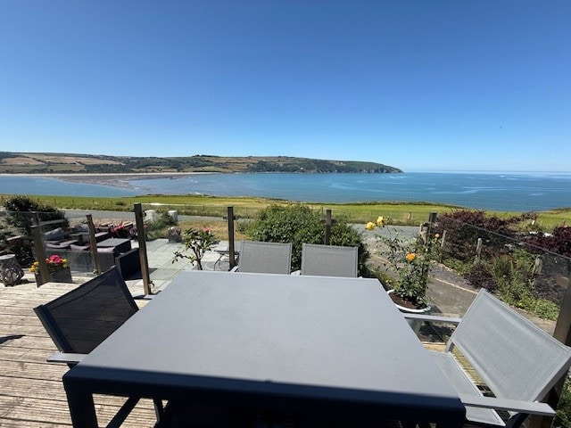 A spacious outdoor dining area is showcased with a rectangular table surrounded by six grey chairs. In the background, panoramic views of the coast and lush greenery are visible, framed by a clear blue sky.