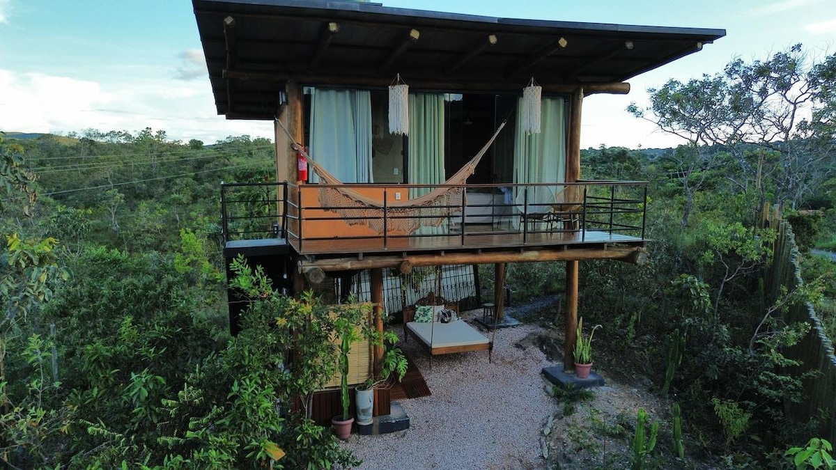 A rustic bungalow is elevated on stilts, featuring a spacious balcony with a hammock and comfortable seating. Surrounding greenery complements the structure, while large glass windows allow for scenic views of nature. The entry area is adorned with potted plants, adding to the tranquil atmosphere.