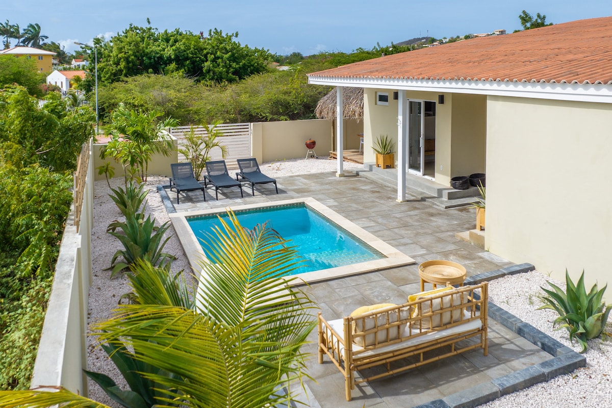 A private pool surrounded by a stone patio is framed by lush greenery. Comfortable lounge chairs are positioned beside the water, while a shaded seating area offers a cozy spot for relaxation. The exterior of the villa is accessible and blends seamlessly into the natural surroundings.