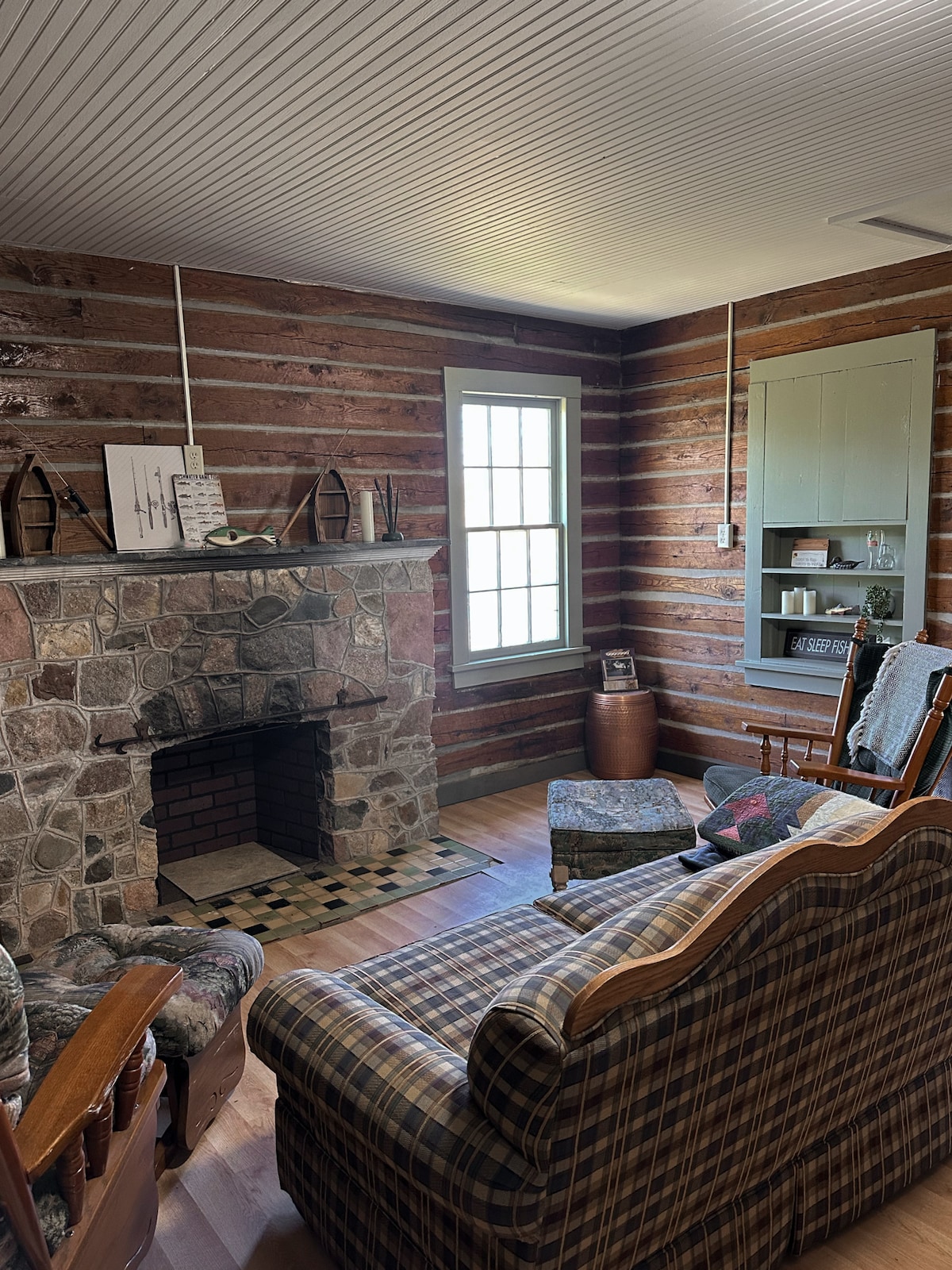 A rustic living area features log cabin walls and hardwood flooring. A stone fireplace sits at one end, complemented by a wooden rocking chair and plaid sofa, providing a cozy seating arrangement. Natural light filters through a large window, highlighting the simple, inviting decor.