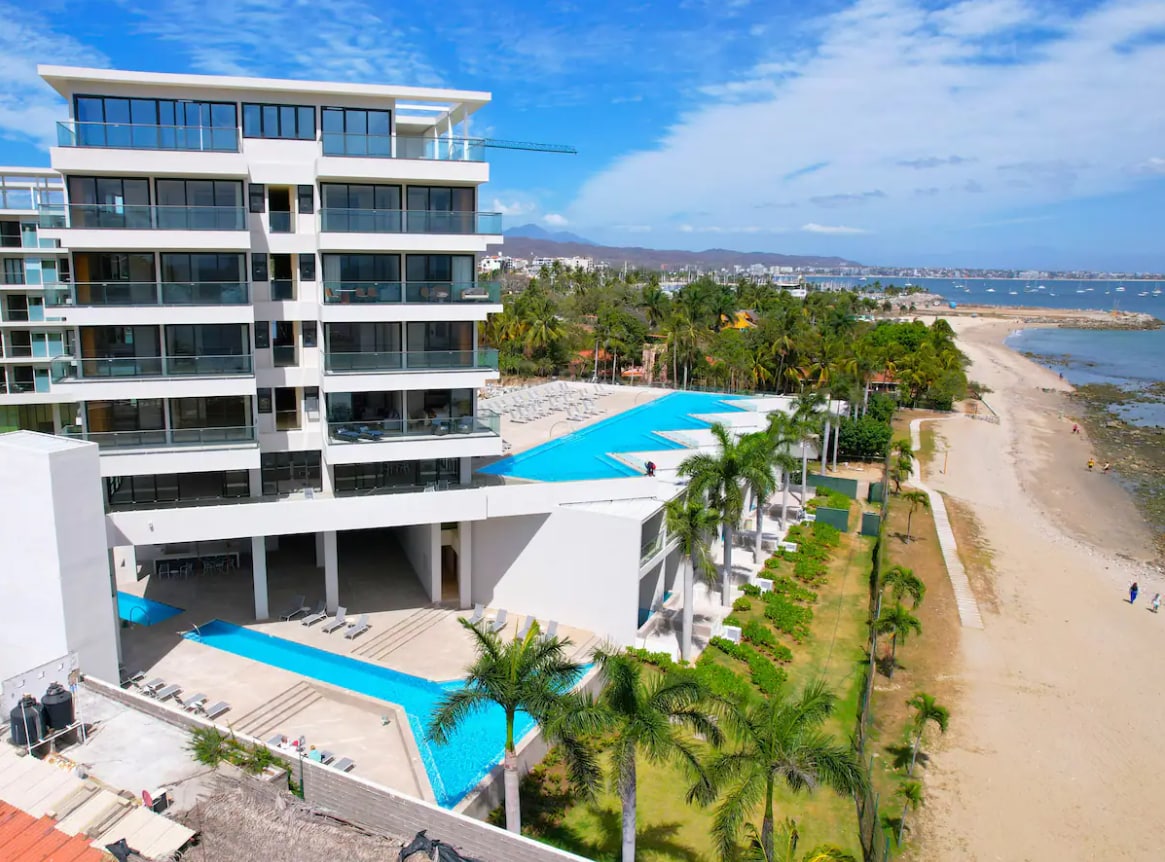The image showcases a modern beachfront condo with three expansive infinity pools designed for relaxation. Lush greenery surrounds the property, which is positioned near a sandy shoreline. A clear blue sky complements the serene atmosphere, enhancing the seaside view.