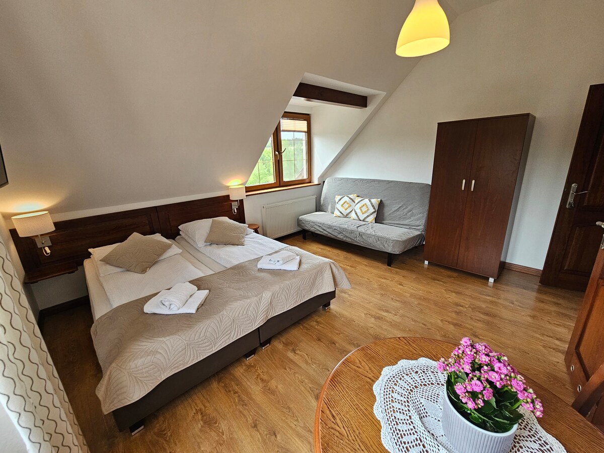 A bright room features a spacious bed configuration with soft bedding, complemented by a modern sofa. Natural light streams through a window, enhancing the relaxing atmosphere. A wardrobe provides ample storage, while a small table adorned with a flower pot adds a touch of charm.