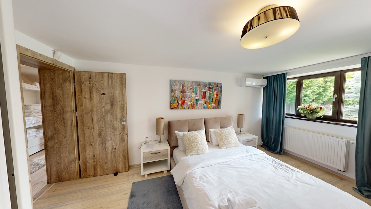 A serene bedroom features a spacious bed with soft linens and decorative pillows. Bedside tables with lamps provide subtle lighting, while a colorful art piece adorns the wall above. Large curtains frame the window, allowing natural light to illuminate the room.