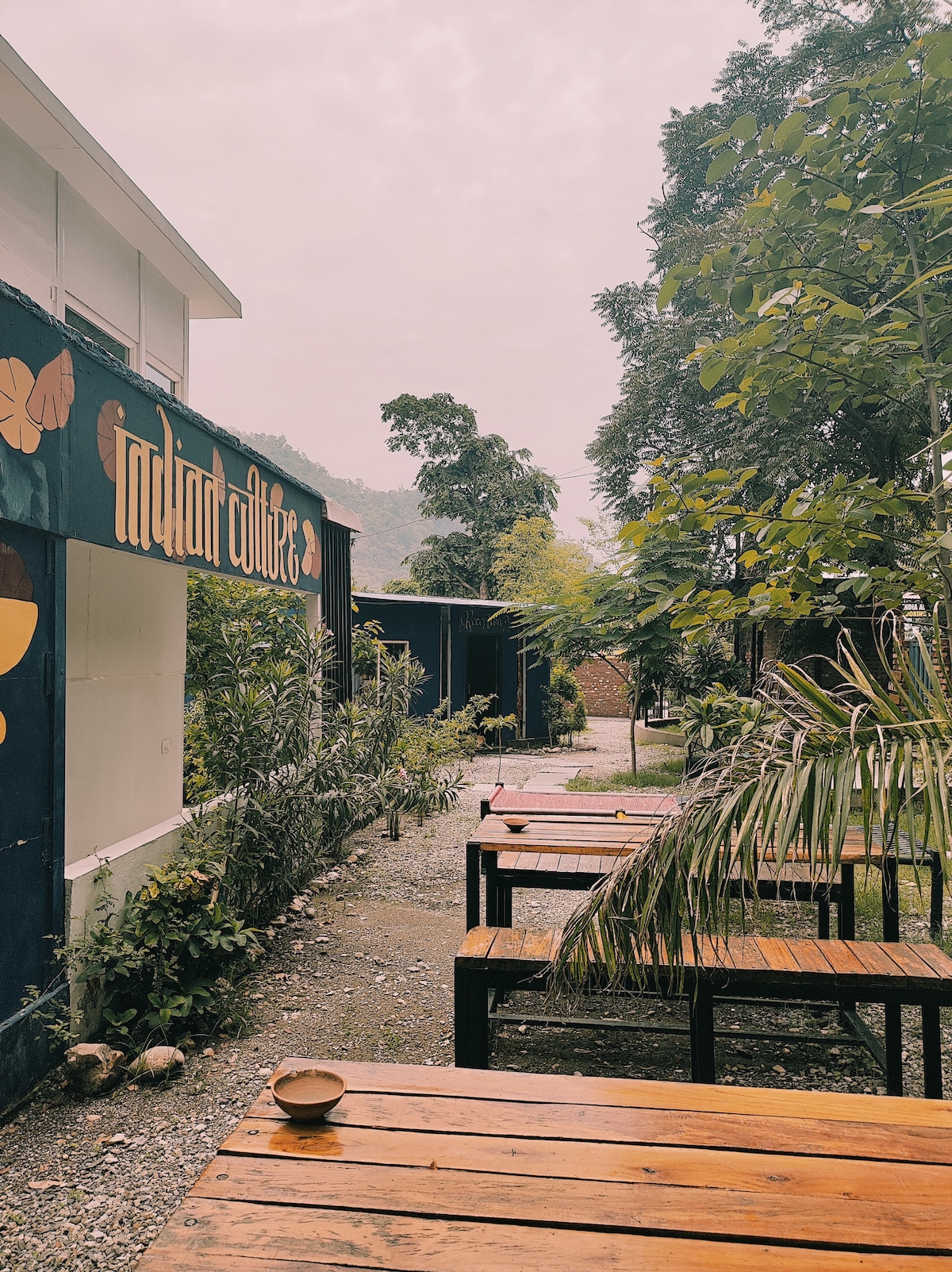 An outdoor area is shown with several wooden tables arranged along a gravel pathway. Lush greenery lines the sides, with a vibrant sign featuring butterflies attracting attention. The scene is set against a backdrop of distant hills, creating a serene environment.