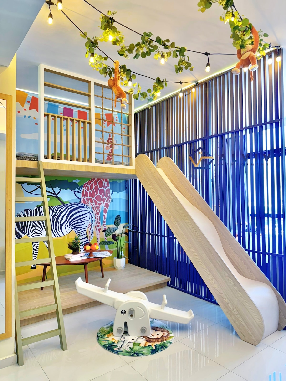 A playful children's area is accentuated by a wooden slide and climbing structure. Colorful wall art featuring animals creates a vibrant backdrop. A small table is positioned in front, accompanied by a green seat for comfortable playtime. String lights add a whimsical touch to the space.