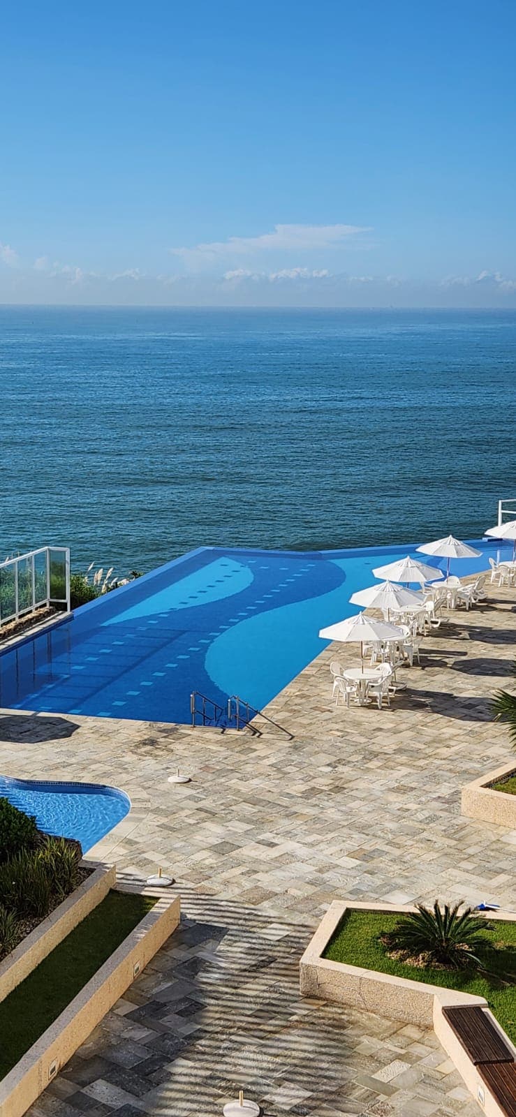 An infinity pool extends toward the ocean, featuring a gentle curve and steps leading into the water. Surrounding the pool, white sun umbrellas offer shade on the stone terrace, complemented by well-manicured greenery and a breathtaking view of the sea under a clear blue sky.