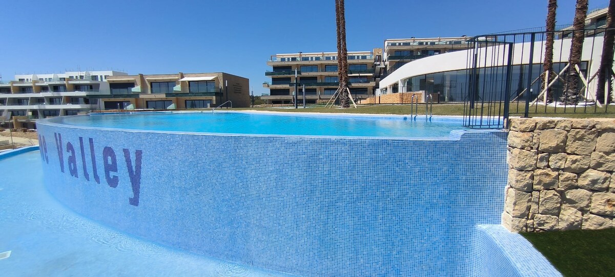 An infinity pool is featured, with a smooth blue mosaic surface reflecting the sky. The pool is enclosed by a stone wall and a metal fence, providing a clear view of the surrounding greenery and buildings. Palm trees add to the serene atmosphere.