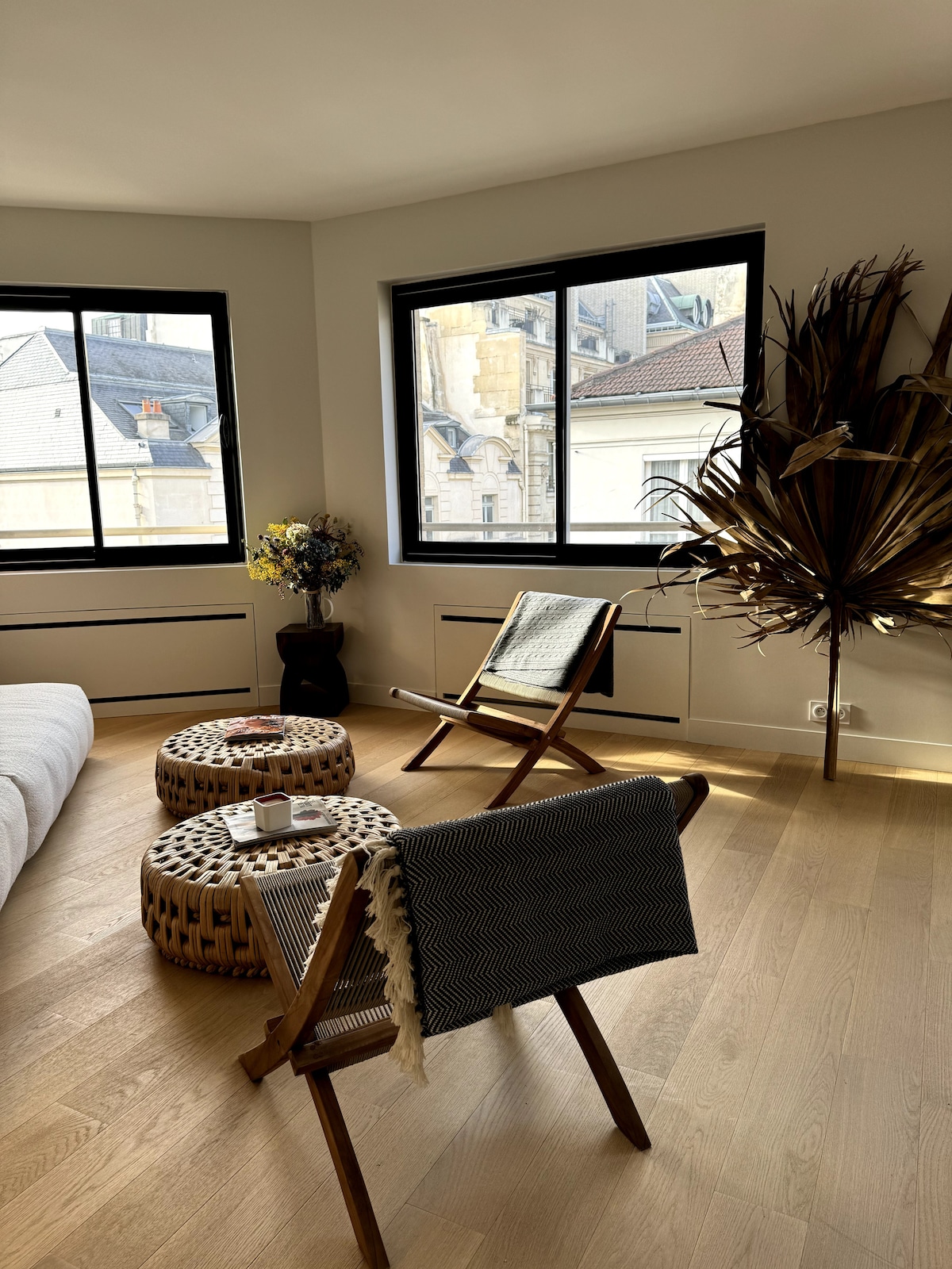 A bright living area features large windows that allow natural light to fill the room. Two wooden chairs are positioned beside intricately designed tables. A tall decorative plant stands in the corner, complemented by a small bouquet of flowers on a table, enhancing the inviting ambiance.