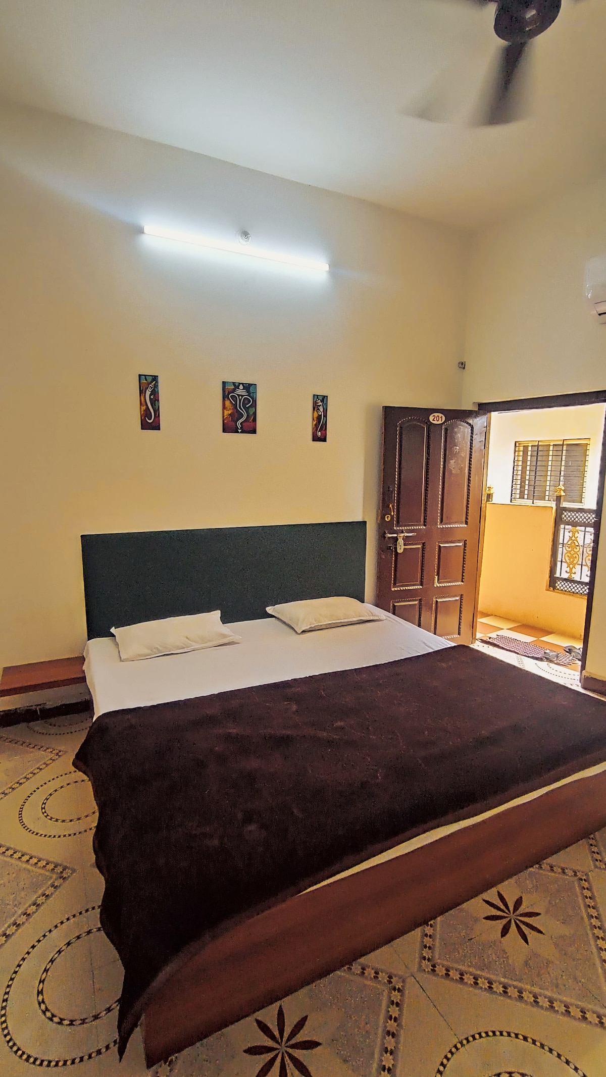 A bedroom features a spacious bed with a dark blanket, complemented by soft pillows. Minimalist artwork adorns the wall above the bed. Natural light filters in from a nearby window, while the room is illuminated by overhead lighting.