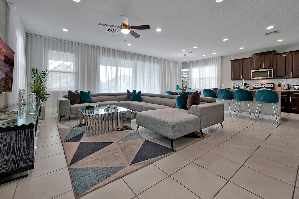 A spacious open-plan living area features a large sectional sofa complemented by accent pillows. A modern coffee table sits atop a geometric area rug, while a wall-mounted Smart TV is visible in the background. Large windows draped with light curtains allow ample natural light to fill the room.